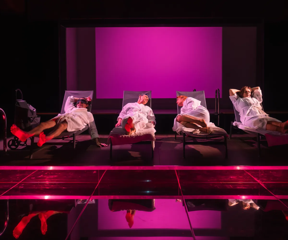 Scena ze spektaklu „Pływalnia” w Teatrze Jaracza. Czworo aktorów w białych szlafrokach leży obok siebie na leżakach basenowych ustawionych w głębi sceny. Całość tonie w intensywnym, różowo-fioletowym świetle reflektorów. Na pierwszym planie znajduje się gładka, ciemna podłoga, w której odbijają się postacie i zarysy leżaków, a po lewej stronie widać metalową drabinkę basenową.