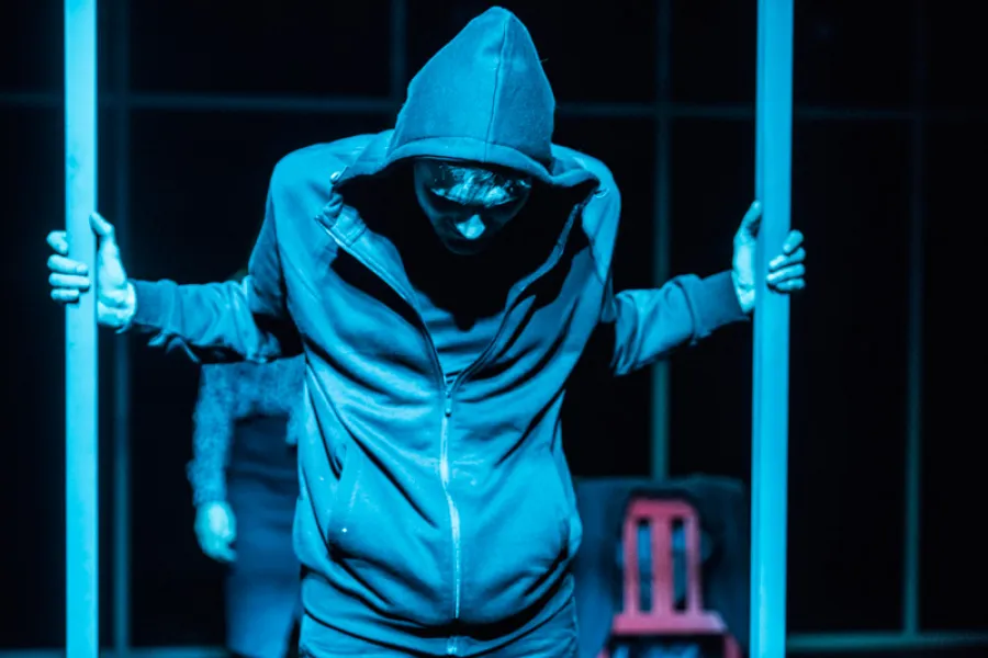 Mężczyzna w bluzie z kapturem stoi na scenie w niebieskim oświetleniu, trzymając się rękami pionowych słupów i patrząc w dół. W tle widoczny jest zarys postaci oraz czerwone krzesło.