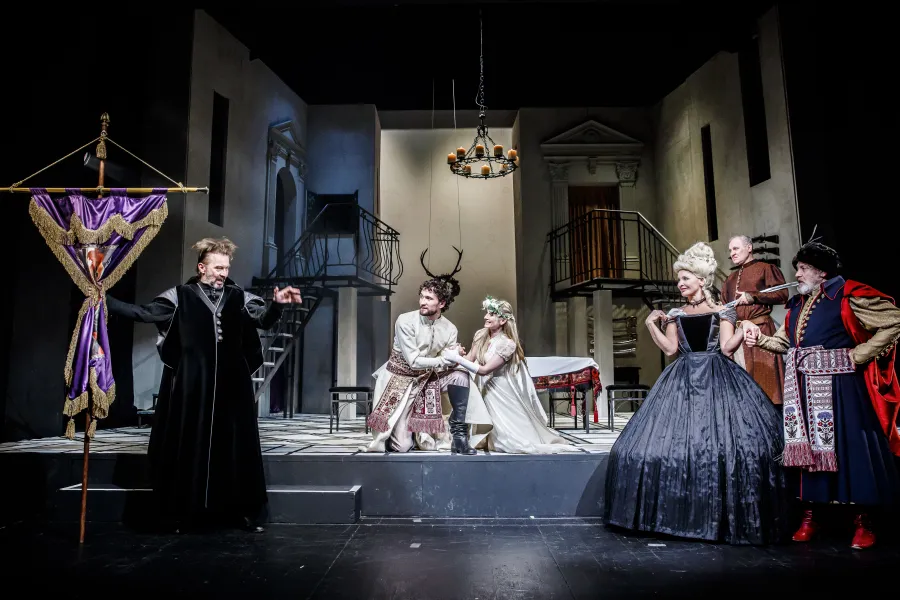 Grupa aktorów w strojach z epoki występuje na scenie teatralnej, gdzie para w jasnych strojach klęczy, trzymając się za ręce. Po lewej stronie mężczyzna w czarnej szacie prezentuje fioletowy sztandar, a po prawej para szlachecka w błękitnym kontuszu i czarnej sukni przygląda się ceremonii.
