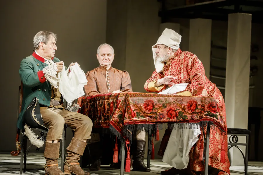 Trzej mężczyźni w historycznych strojach siedzą przy stole nakrytym wzorzystym obrusem. Po lewej mężczyzna w zielonym mundurze ogląda białą tkaninę, a po prawej brodaty aktor w czerwonym, bogato zdobionym żupanie i czapce żywo z nim rozmawia.