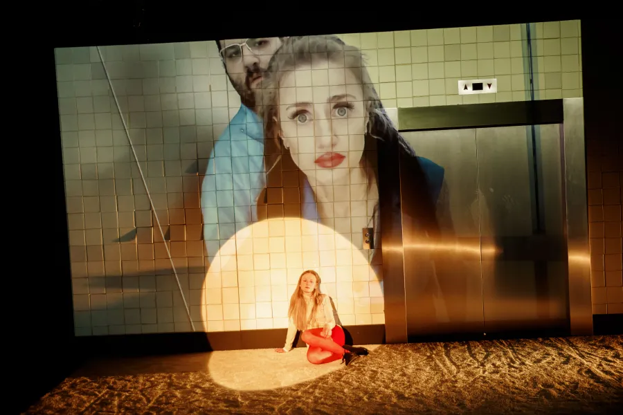 Na scenie teatralnej siedząca w okręgu światła kobieta z jasnymi włosami znajduje się na tle wielkoformatowej projekcji twarzy innych aktorów. Obok projekcji widoczne są zamknięte drzwi windy i kafelkowa ściana.