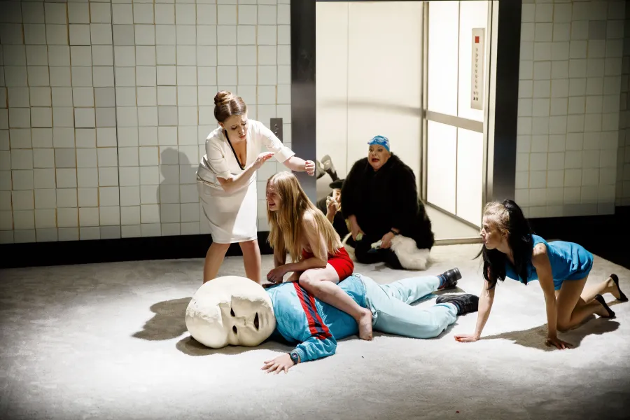 Na scenie teatralnej kobieta w białej sukience stoi nad płaczącą dziewczyną w czerwonej sukience, która siedzi na leżącym na ziemi mężczyźnie z dużą, białą, sztuczną głową. Obok widać kobietę w niebieskim stroju i inną postać w futrze.