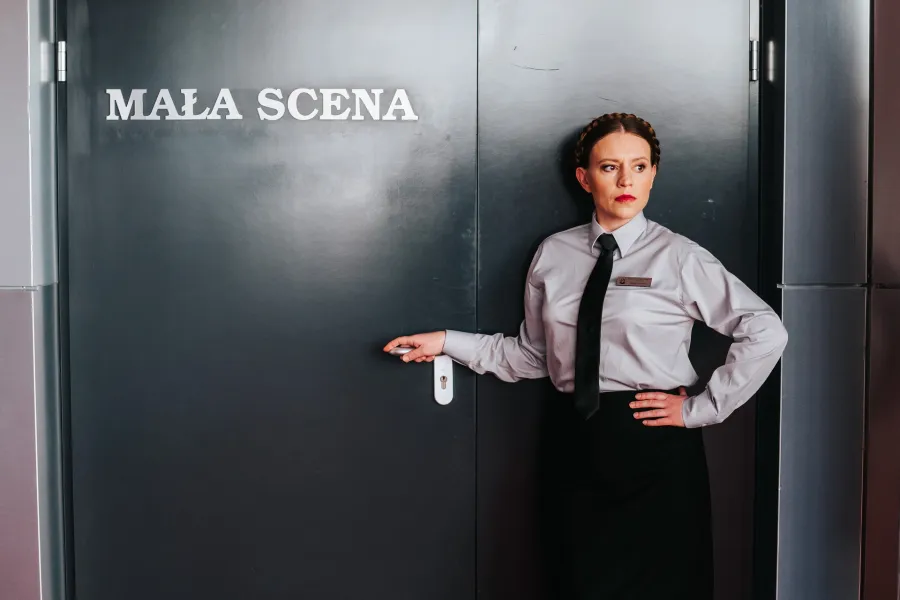 Kobieta w szarej koszuli, czarnym krawacie i z identyfikatorem stoi przy czarnych drzwiach z napisem „MAŁA SCENA”, trzymając dłoń na klamce. Drugą rękę opiera na biodrze i patrzy w bok z poważnym wyrazem twarzy.
