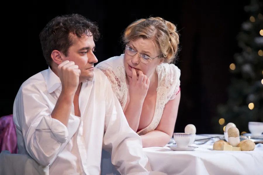 Na scenie teatralnej siedzący przy stole mężczyzna w białej koszuli patrzy w bok z zamyśleniem, podczas gdy stojąca obok niego kobieta w okularach i jasnej bluzce delikatnie gryzie palec, pochylając się w jego stronę. Na nakrytym białym obrusem stole stoją filiżanki i talerze z jajkami oraz pieczywem.