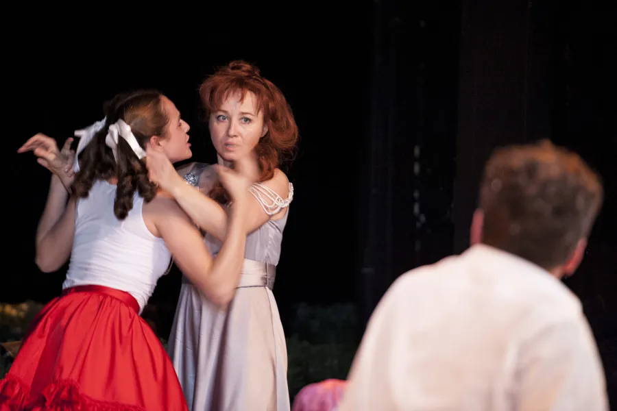 Na scenie teatralnej rudowłosa kobieta w szarej sukni próbuje rozdzielić szarpiące się kobiety, z których jedna ma na sobie czerwoną spódnicę i białe kokardy we włosach. Na pierwszym planie plecami do widza siedzi mężczyzna w białej koszuli, obserwujący tę scenę.
