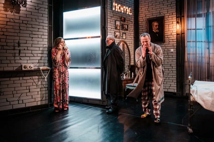 Na scenie teatralnej stylizowanej na pokój stoją trzy postacie wokół łóżka i jasnego panelu świetlnego. Mężczyzna w szlafroku i piżamie w kratkę trzyma ręce przy twarzy z zatroskaną miną, obok niego stoi mężczyzna w ciemnym płaszczu, a po lewej stronie kobieta w długim szlafroku.
