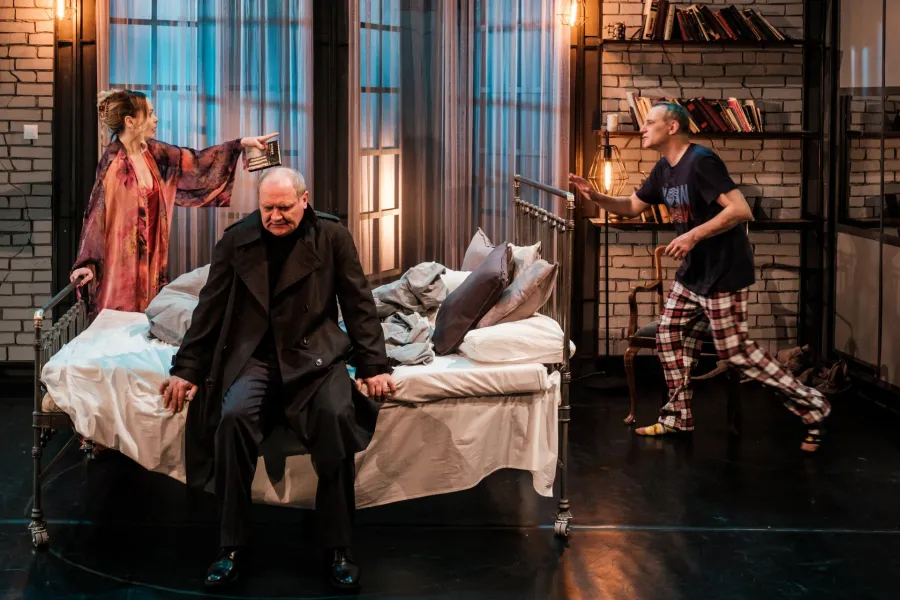 Na scenie teatralnej kobieta w kolorowej narzutce trzyma książkę nad głową siedzącego na łóżku mężczyzny w ciemnym płaszczu. Po prawej stronie, w kierunku łóżka, zmierza mężczyzna w piżamie i ciemnej koszulce.