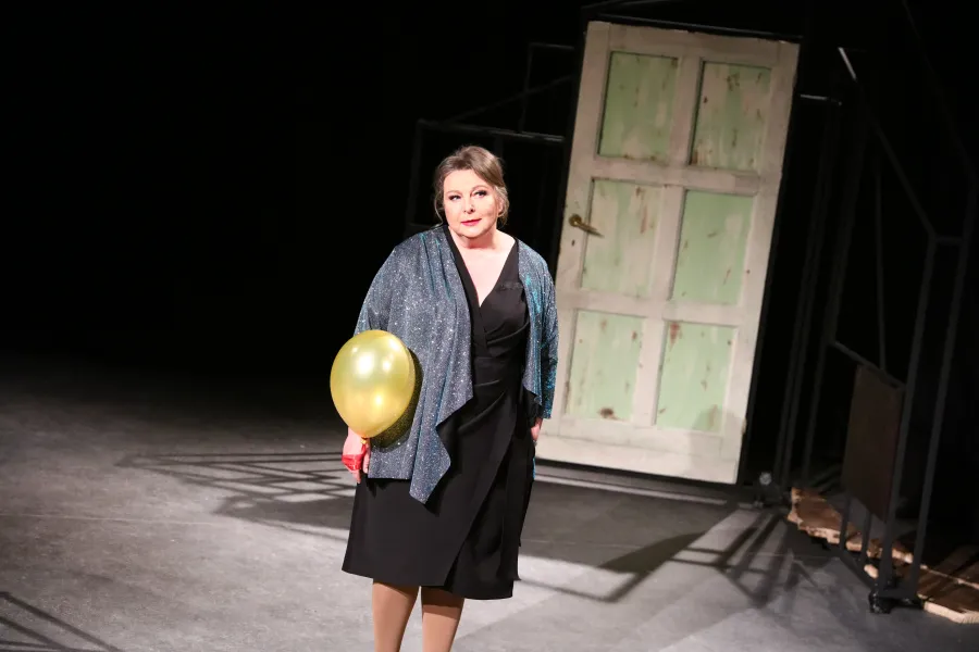 Kobieta w eleganckiej czarnej sukience i błyszczącym narzuconym szalu stoi na scenie teatralnej, trzymając w ręku żółty balonik. W tle widoczne są jasne, stare drzwi osadzone w ciemnej konstrukcji scenograficznej.