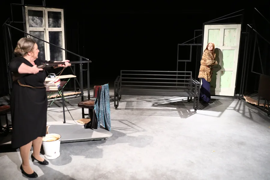 Na scenie teatralnej kobieta w czarnej sukience celuje z długiej broni w stronę drzwi, w których stoi inna kobieta w futrzanym płaszczu. W tle między nimi znajduje się metalowa ławka oraz elementy minimalistycznej scenografii.