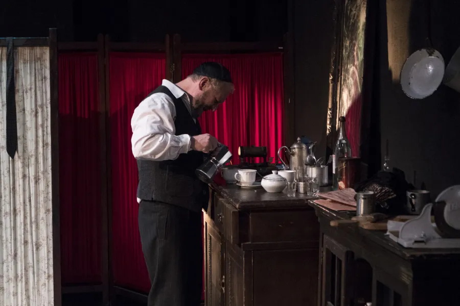 Mężczyzna w białej koszuli, kamizelce i jarmużce stoi pochylony nad komódką w teatralnej scenografii i nalewa płyn z kawiarki do filiżanki. W tle widoczny jest czerwony parawan oraz naczynia na blacie.