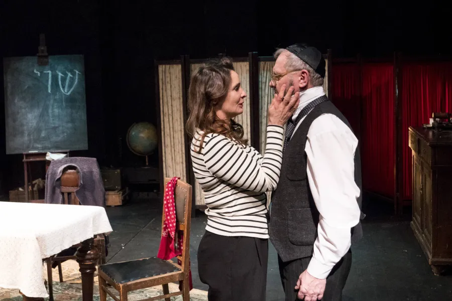 Kobieta w bluzce w paski stoi na scenie teatralnej, czule dotykając twarzy mężczyzny w jarmużce, białej koszuli i ciemnej kamizelce. W tle widoczna jest scenografia pokoju z tablicą, globusem i nakrytym stołem.