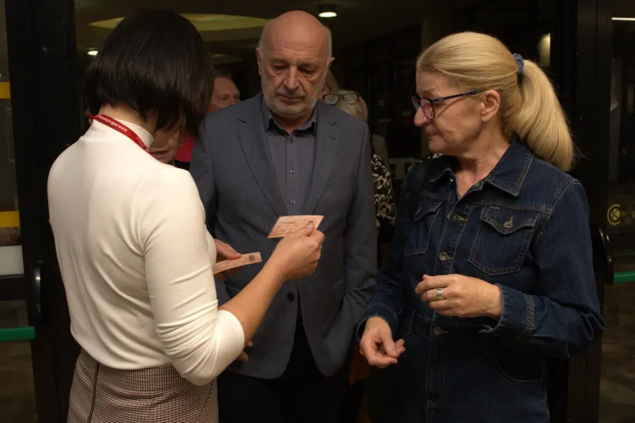 Na zdjęciu jest dwójka widzów - kobieta i mężczyzna, którzy mają sprawdzane bilety przed wejściem na salę teatralną. Bileterka stoi tyłem do aparatu.