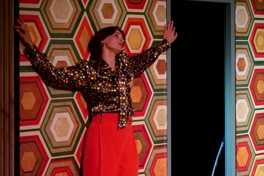 Kobieta w brązowej bluzce w groszki i pomarańczowych spodniach stoi na scenie teatralnej przy ścianie w geometryczne, kolorowe wzory. Opiera się plecami o ścianę z rękami rozłożonymi szeroko po bokach, patrząc w górę.