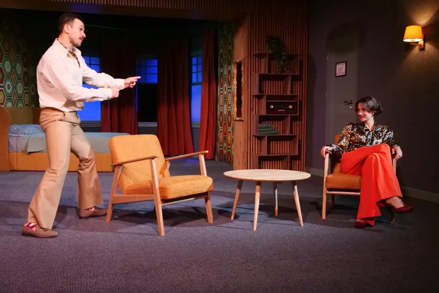 Na scenie teatralnej mężczyzna w jasnej koszuli i spodniach stoi z wyciągniętymi rękami, patrząc na siedzącą w fotelu kobietę w pomarańczowych spodniach i wzorzystej bluzce. W tle widać wystrój pokoju hotelowego z łóżkiem, stolikiem i geometrycznymi tapetami.