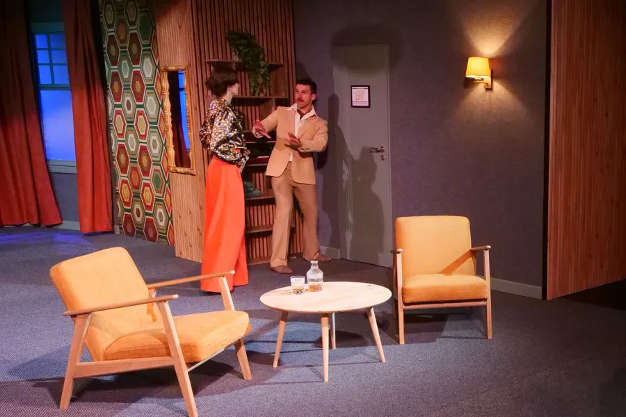 Na scenie teatralnej kobieta w pomarańczowych spodniach i wzorzystej bluzce stoi tyłem, a stojący naprzeciwko niej mężczyzna w jasnym garniturze wykonuje gest otwartymi dłońmi. Sceneria przypomina pokój hotelowy z dwoma żółtymi fotelami, niskim stolikiem i drewnianymi panelami ściennymi.