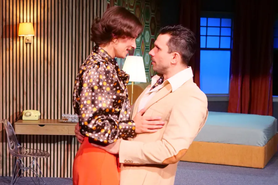 Na scenie teatralnej kobieta w brązowej bluzce w groszki i pomarańczowych spodniach obejmuje stojącego naprzeciwko mężczyznę w jasnym garniturze. W tle widoczny jest wystrój pokoju hotelowego z łóżkiem oraz stolikiem.
