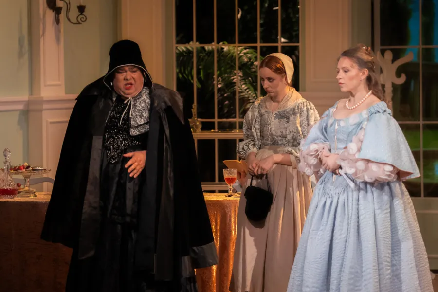 Trójka aktorów w kostiumach z epoki baroku występuje na scenie teatralnej, która przedstawia wnętrze jadalni. Postać po lewej w czarnej pelerynie i kapeluszu mówi z zaangażowaniem, podczas gdy dwie kobiety w sukniach – jednej wzorzystej z czepkiem, drugiej błękitnej z perłami – słuchają jej uważnie.