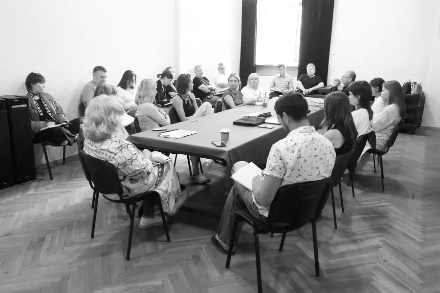 pierwsza próba spektaklu „Czas na wielki numer". Zdjęcie zostało zrobione na sali prób. Po środku widać duży stół nakryty suknem, na stole znajdują się egzemplarze scenariusza, notesy oraz kubki. Przy stole siedzą realizatorzy i aktorzy występujący w spektaklu. Wokół stołu na krzesłach siedzą pracownicy teatru. Na zdjęcie nałożony jest czarno-biały filtr. Fot. Marcin Nowak