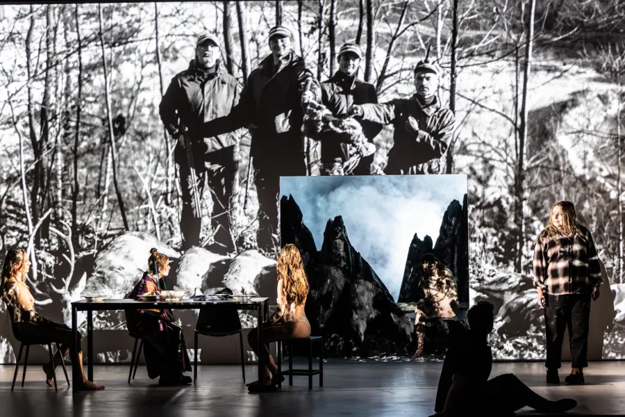Na Dużej Scenie Teatru Jaracza widać długi stół przy którym siedzą dwie tancerki i aktorka Sara Lityńska, patrzą się na wyświetlane na ścianie zdjęcie myśliwych, w ścianie jest kwadratowa dziura przez którą widać góry. Obok dziury stoi aktorka Milena Lisiecka. Autorką zdjęcia jest Natalia Kabanow