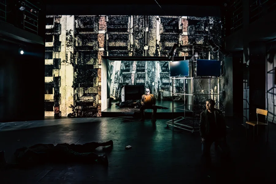 Scena teatralna przedstawia ciemne, surowe wnętrze z projekcją zniszczonego budynku w tle. W głębi siedzi postać, na pierwszym planie po prawej klęczy mężczyzna, a na podłodze leżą inne osoby.