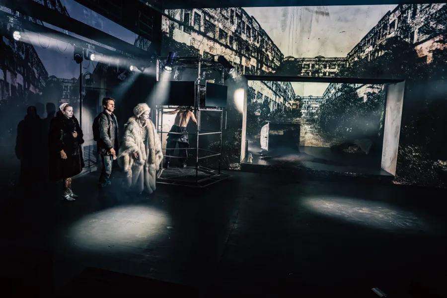 Na scenie teatralnej oświetlonej reflektorami stoi grupa aktorów, w tym postać w puszystym futrze. W tle widoczna jest duża projekcja przedstawiająca zniszczone budynki oraz metalowa konstrukcja sceniczna.