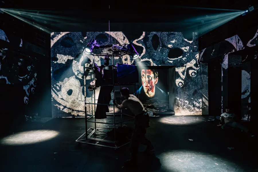 Na scenie teatralnej widać półnagiego mężczyznę stojącego obok metalowej konstrukcji z reflektorami. W tle wyświetlana jest duża, abstrakcyjna projekcja oraz twarz człowieka na mniejszym ekranie.
