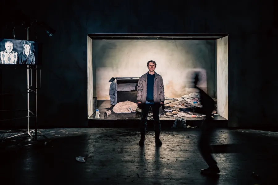 Na scenie teatralnej, w prostokątnym oknie, stoi mężczyzna w kurtce, za którym widać śmieci i przewrócony kontener. Po lewej stronie na stojaku znajduje się monitor wyświetlający czarno-białe zdjęcie dwóch osób, a po prawej mija go szybko rozmazana postać.