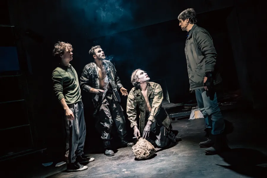 Na scenie teatralnej grupa czterech mężczyzn znajduje się w ciemnej przestrzeni oświetlonej punktowym światłem. Dwóch z nich ma pomalowane na biało twarze, jeden klęczy przy kamieniu, a stojący po prawej stronie mężczyzna w kurtce wpatruje się w pozostałych.