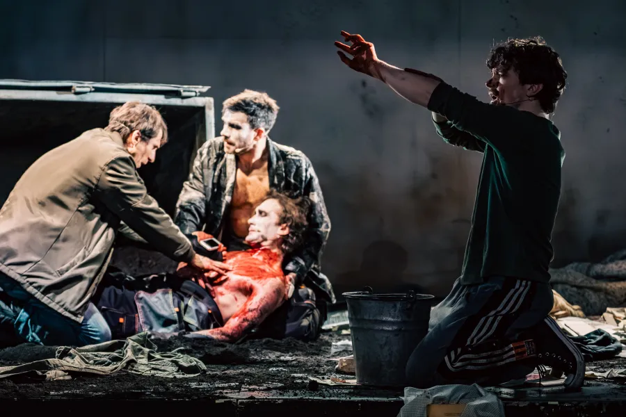 Na scenie teatralnej grupa postaci pochyla się nad leżącym, posiniaczonym i zakrwawionym człowiekiem. Po prawej stronie klęczy mężczyzna z uniesionymi rękami.