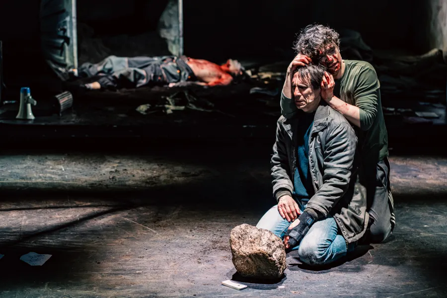 Na scenie teatralnej klęczy dwóch mężczyzn, z których jeden, cały we krwi, przytrzymuje głowę siedzącego przed nim człowieka. W tle na podłodze leży nieruchomo inna postać w dramatycznej scenerii.