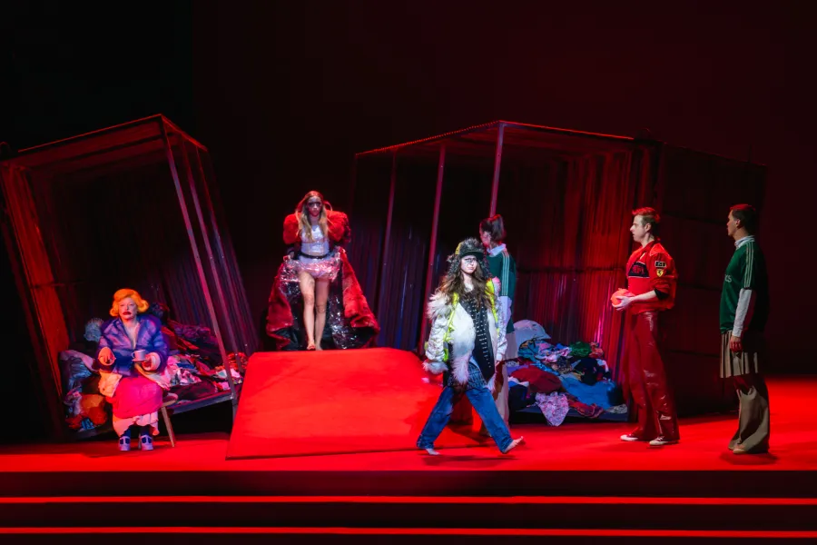 Teatralna scena w czerwonym oświetleniu z performerami w nietypowych strojach, stojącymi wśród metalowych konstrukcji i stosów ubrań. Centralnie postać w bieliźnie i czerwonej płachcie oraz inna w futrze i dżinsach, otoczone osobami w błękitnym stroju, czerwonej kurtce i zielonej koszulce.