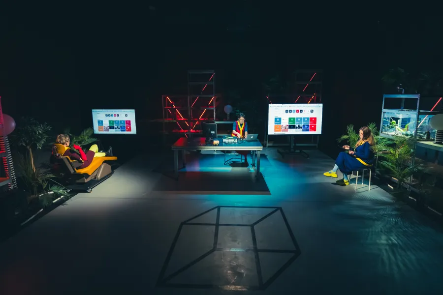 Trzy osoby znajdują się na nowoczesnej, mrocznej scenie teatralnej, podzielonej na osobne stanowiska pracy z ekranami i czerwonokrzemowym oświetleniem w tle. Jedna osoba leży na rozkładanym fotelu po lewej stronie, druga pracuj przy biurku pośrodku, a trzecia siedzi na krześle po prawej stronie w otoczeniu roślin i regałów.