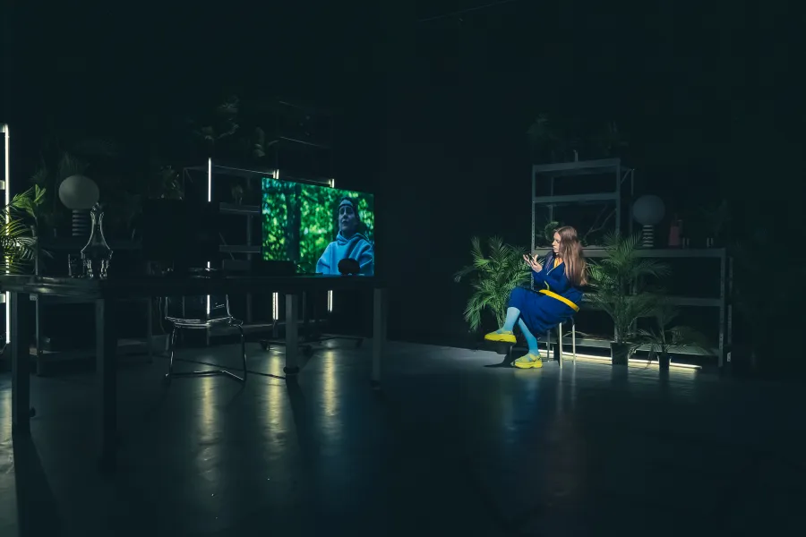 Kobieta w niebiesko-żółtej sukni siedzi na krześle na zaciemnionej scenie teatralnej i korzysta ze smartfona w otoczeniu roślin. Po lewej stronie widać duży ekran z wyświetlaną twarzą postaci w zielonym otoczeniu oraz zarys biurka ze stanowiskiem pracy.