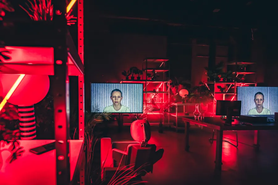 Mroczna, laboratoryjna przestrzeń sceniczna skąpana jest w intensywnym, czerwonym świetle nieonowym. Na dwóch dużych monitorach rozstawionych wśród regałów z roślinami wyświetlany jest ten sam portret kobiety.
