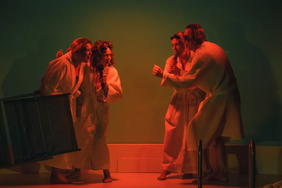 Scena ze spektaklu „Pływalnia” w Teatrze Jaracza. Czworo aktorów ubranych w białe szlafroki kąpielowe stoi na scenie, podzielonych na dwie ekspresyjne pary stojące naprzeciw siebie. Dwie osoby trzymają mikrofony, a wszyscy mają dynamiczne, pełne emocji miny, jakby śpiewali lub krzyczeli. Całość oświetlona jest intensywnym, kontrastowym światłem – czerwonym od dołu i zielonym w tle, co nadaje scenie dramatyczny charakter. Po lewej stronie widoczny jest przewrócony leżak, a po prawej fragment drabinki