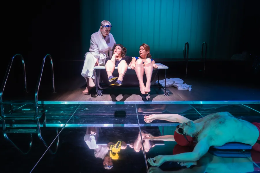 Scena ze spektaklu „Pływalnia” w Teatrze Jaracza, podzielona na dwa plany. Na podwyższeniu w głębi sceny trzy aktorki – jedna w białym szlafroku, dwie w strojach kąpielowych – siedzą ściśnięte w czarnej wannie na kółkach i rozmawiają. Na pierwszym planie, na błyszczącej, lustrzanej podłodze imitującej taflę wody, leży na plecach aktor w czerwonych kąpielówkach i czepku. Całość oświetlona jest chłodnym, turkusowym światłem, a z boku widoczne są drabinki basenowe.