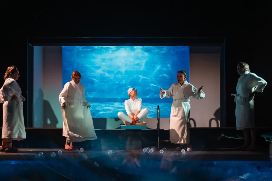 Scena ze spektaklu „Pływalnia” w Teatrze Jaracza. W centrum podwyższenia siedzi aktorka w białym stroju i czepku, otoczona przez czworo innych aktorów w białych szlafrokach, którzy żywo gestykulują. W tle widać duży ekran z podwodną, błękitną grafiką basenu, a na pierwszym planie rozsypane, małe kulki.