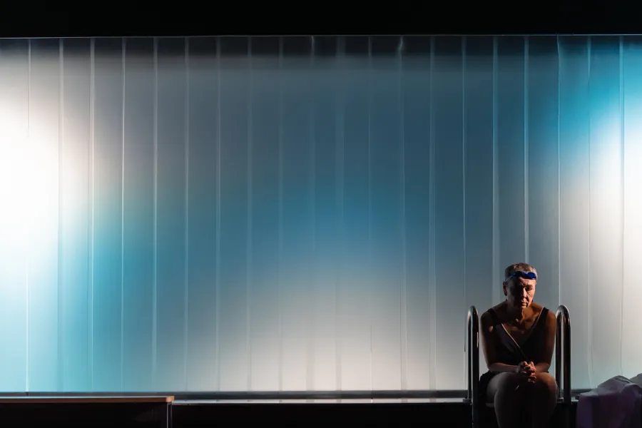 Scena ze spektaklu „Pływalnia” w Teatrze Jaracza. W prawym dolnym rogu kadru siedzi samotna aktorka w ciemnym stroju kąpielowym i goglach na głowie, mając splątane dłonie i opuszczony wzrok. Tło zdjęcia stanowi duża, minimalistyczna, pionowo prążkowana ściana oświetlona chłodnym, błękitno-białym światłem reflektorów, co tworzy atmosferę pustki i melancholii. Obok kobiety widoczna jest metalowa drabinka basenowa.