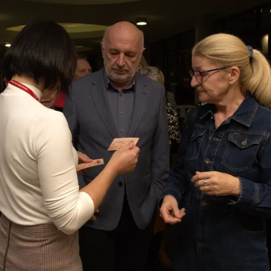 Na zdjęciu jest dwójka widzów - kobieta i mężczyzna, którzy mają sprawdzane bilety przed wejściem na salę teatralną. Bileterka stoi tyłem do aparatu. 