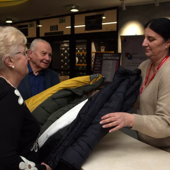 Zdjęcie przedstawia dwójkę widzów oddających do szatni swoje kurtki, które odbiera szatniarka. 