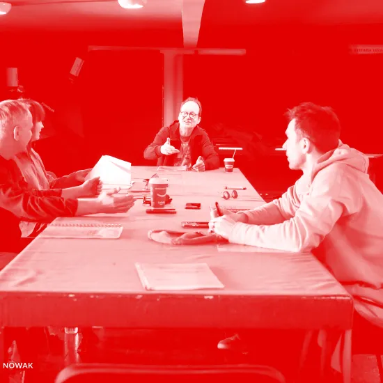 zdjęcie przedstawia pierwszą próbę spektaklu "Morderstwo w hotelu". Przy stole siedzą aktorzy, inspicjentka i reżyser. Po lewej stronie siedzą aktor Mariusz Witkowski i inspicjentka Marta Baraszkiewicz, po środku na szczycie stołu siedzi reżyser Jacek Bończyk, który coś tłumaczy. Po prawej stronie siedzą aktorzy Natalia Klepacka i Krzysztof Wach. Na stole znajdują się egzeplarze tekstu. Na zdjęcie nałożony został czerwony filtr. 