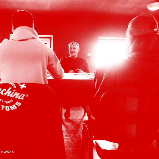 Zdjęcie przedstawia pierwszą próbę spektaklu „Morderstwo w hotelu". Tyłem do obiektywu siedzą przy stole aktorzy Krzysztof Wach i Natalia Klepacka (ona w czapce na głowie). Przodem do obiektywu, po drugiej stronie stołu siedzi aktor Mariusz Witkowski. Za nim znajdują się dwa okna, przez które przebija mocne światło słoneczne. Na zdjęcie został nałożony czerwony filtr. 