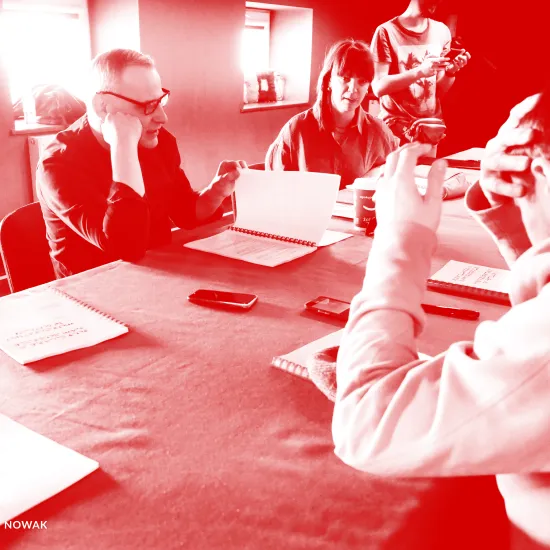 zdjęcie przedstawia pierwszą próbę spektaklu „Morderstwo w hotelu". Po lewej stronie stołu siedzi aktor Mariusz Witkowski czytający egzemplarz sztuki i przerzuca jego kartkę. Obok siedzi inspicjentka Marta Baraszkiewicz. W tle widać pracownika teatru Marcina Majczaka. Po prawej stronie blisko obiektywu siedzi aktor Krzystzof Wach zasłaniający prawą ręką swoją głowę. Lewą rekę ma uniesioną. Na zdjęcie został nałożony czerwony filtr. 