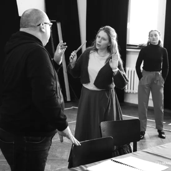 zdjęcie przedstawia próbę spektaklu „Świętoszek". Zostało zrobione na dużej sali prób w Teatrze Jaracza w Łodzi. Tyłem do aparatu stoi aktor Paweł Tucholski. Na przeciwko niego stoi aktorka Matylda Paszczenko, w ręku trzyma zwinięty wachlarz. W tle stoi aktorka Iwona Dróżdż.