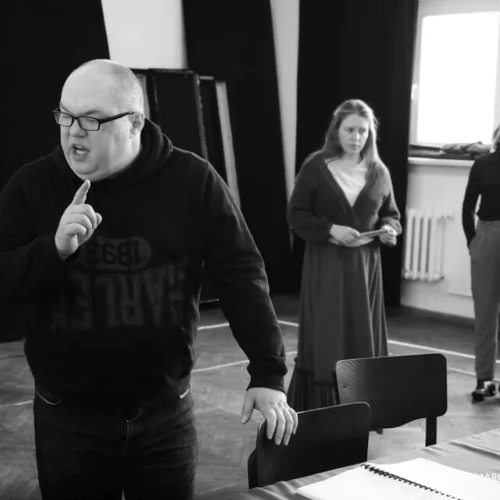 zdjęcie przedstawia próbę spektaklu „Świętoszek". Zostało zrobione na dużej sali prób w Teatrze Jaracza w Łodzi. Przodem do aparatu stoi Paweł Tucholski. Prawą dłoń ma uniesioną pokazując palec wskazujący. W tle stoją aktorki Matylda Paszczenko i Iwona Dróżdż