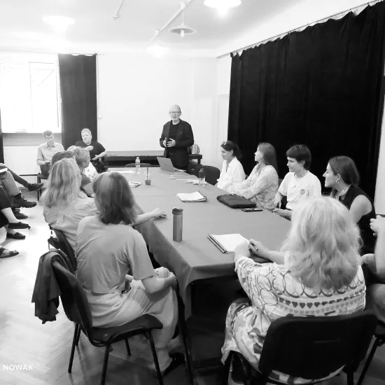 pierwsza próba spektaklu „Czas na wielki numer". Zdjęcie zostało zrobione na sali prób. Po środku widać duży stół nakryty suknem, na stole znajdują się egzemplarze scenariusza, notesy oraz kubki. Przy stole siedzą realizatorzy i aktorzy występujący w spektaklu. Na końcu stołu po prawej stronie stoi dyrektor teatru Paweł Łysak. Wokół stołu na krzesłach siedzą pracownicy teatru. Na zdjęcie nałożony jest czarno-biały filtr. Fot. Marcin Nowak