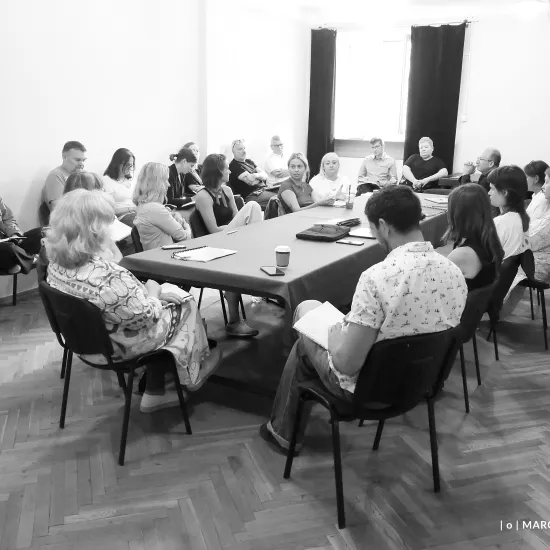 pierwsza próba spektaklu „Czas na wielki numer". Zdjęcie zostało zrobione na sali prób. Po środku widać duży stół nakryty suknem, na stole znajdują się egzemplarze scenariusza, notesy oraz kubki. Przy stole siedzą realizatorzy i aktorzy występujący w spektaklu. Wokół stołu na krzesłach siedzą pracownicy teatru. Na zdjęcie nałożony jest czarno-biały filtr. Fot. Marcin Nowak